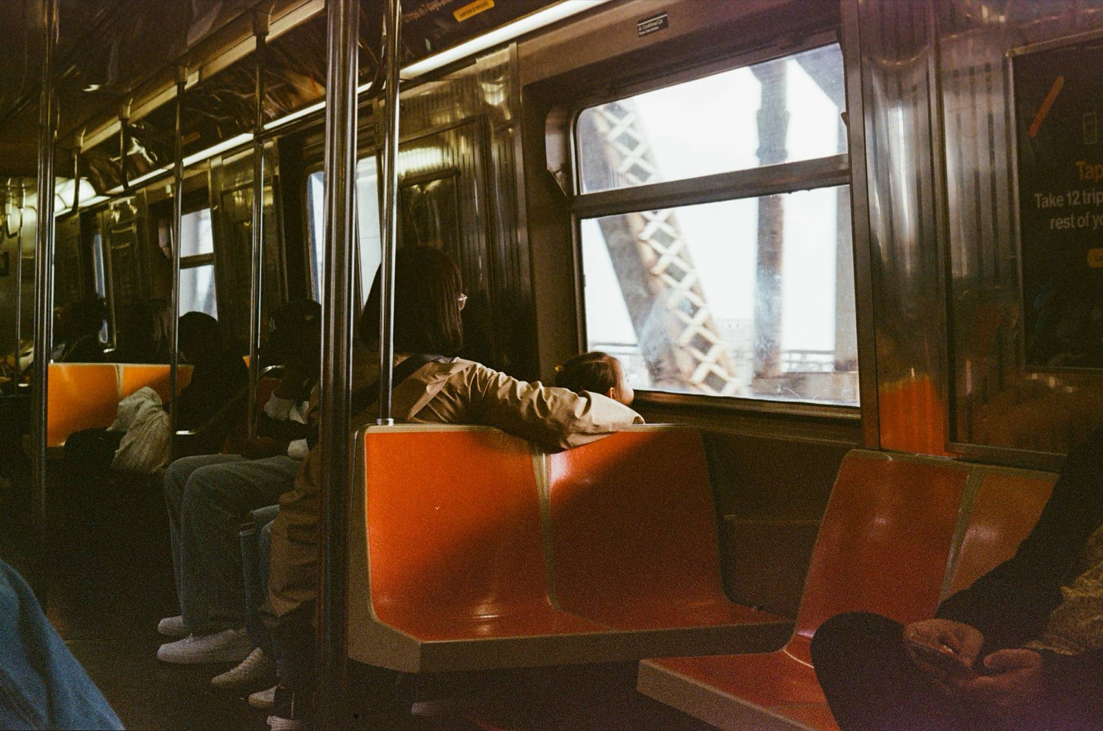Riders on an elevated New York subway car, a child leaning on the window ledge looking out at the bridge